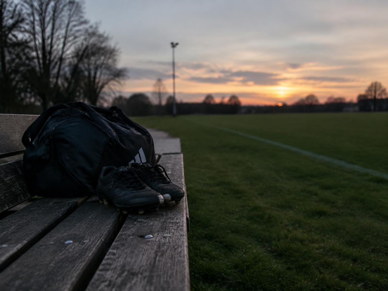 A football kit bag and boots on an empty bench beside a pitch at dusk