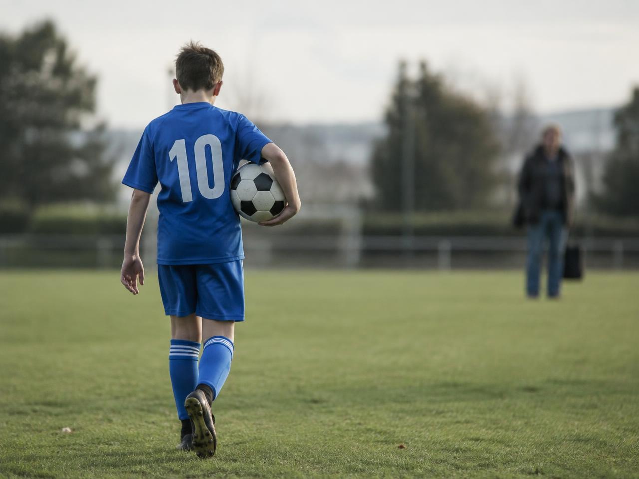 A young footballer walking onto a pitch carrying a ball